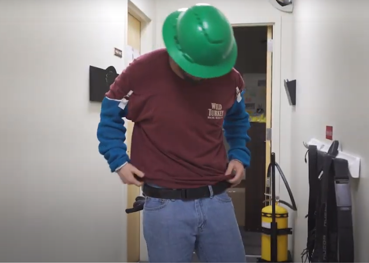 A Caucasian person with their head down and face not visible to camera, wearing a green hard hat, a maroon T-shirt, blue safety sleeves with white buckles at the top, denim blue jeans, and a black Modjoul SmartBelt through the loops of their jeans. The person is standing in a white hallways with two light brown wooden doors and a yellow industrial canister behind them, and a white wall hanger holding several SmartBelts on the wall next to the person.